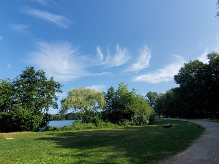 Scenic park with lush green lawn, trees along a waterway, winding path, and blue sky with wispy clouds